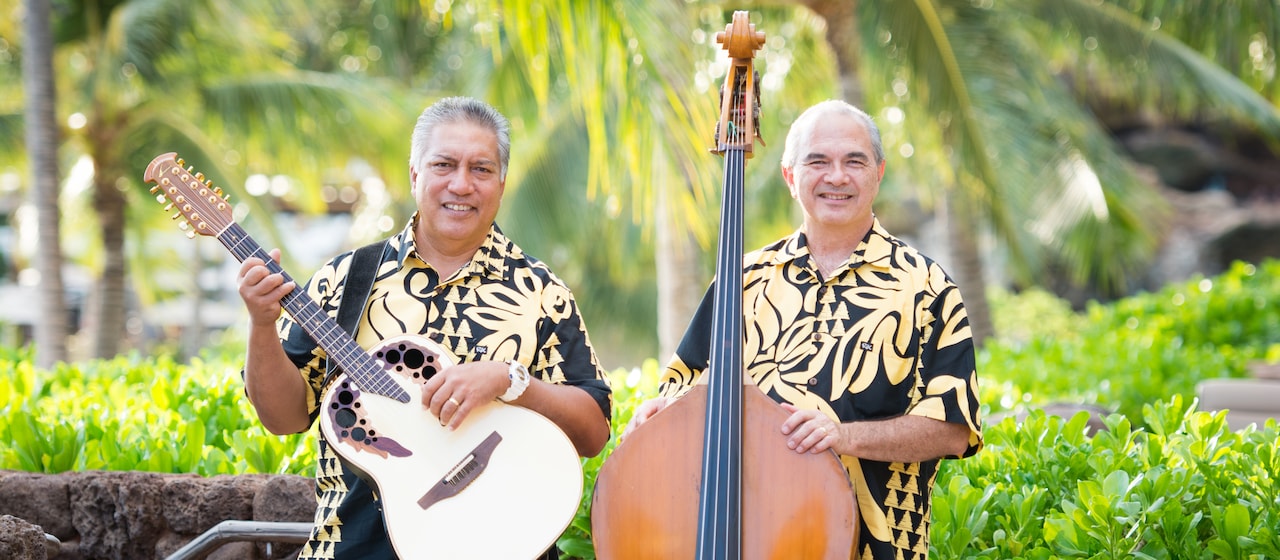 Musical duo Hema Pa’a musicians Baba Alimoot holds his guitar next to Chris Kamaka, who stands next to his stand-up bass wearing matching Aloha shirts